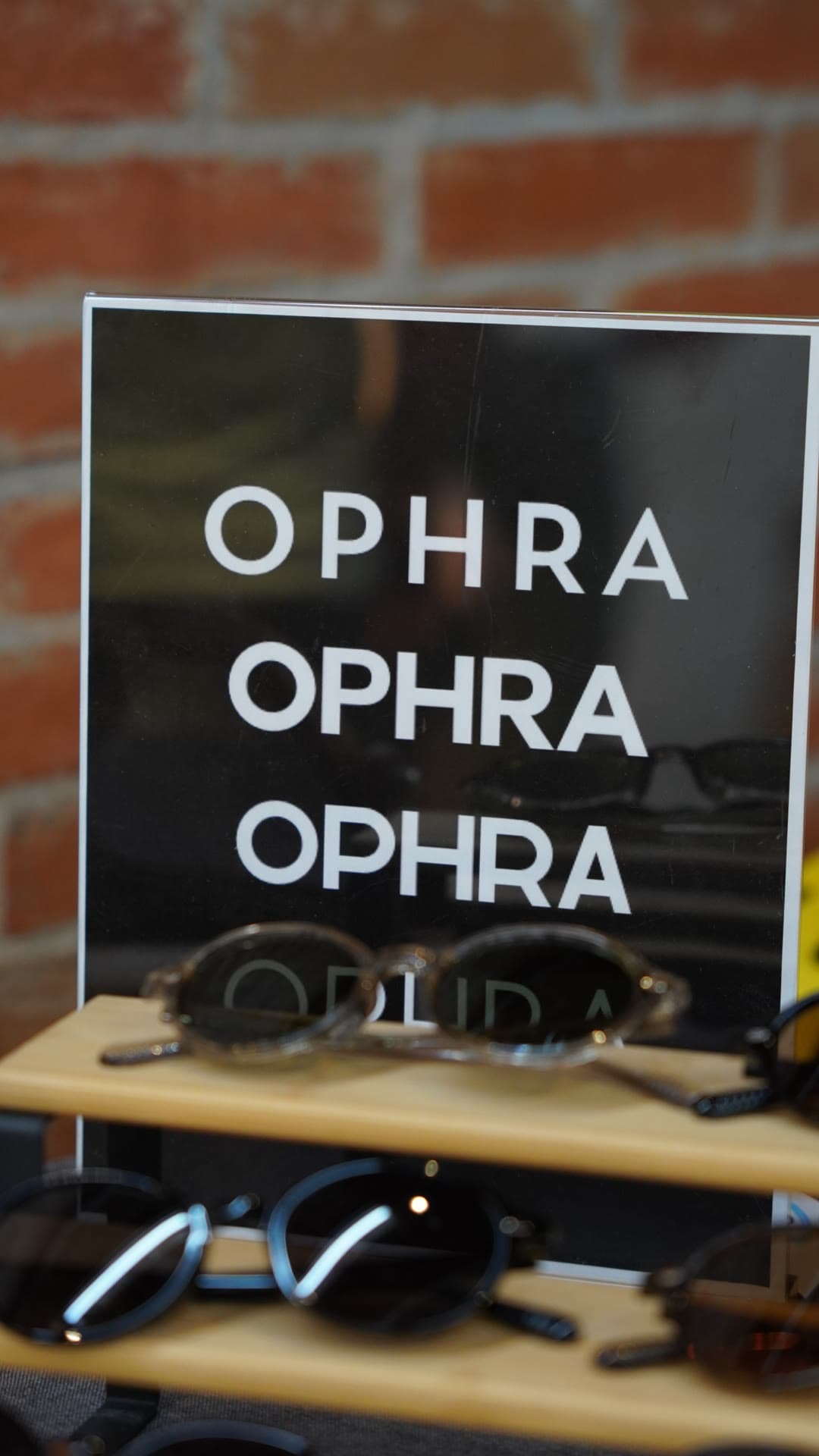 OPHRA display sign and sunglasses on a wooden shelf against a brick wall